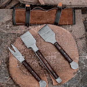 Puede incluir: Un juego de tres utensilios para asar de acero de Damasco con mangos de madera oscura y un estuche de transporte de cuero. Las herramientas incluyen un tenedor y dos espátulas, expuestas sobre una tabla de cortar de madera. El estuche es marrón con correas negras.