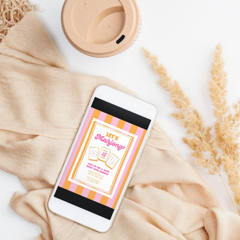 K&ouml;nnte beinhalten: Ein Smartphone zeigt eine rosa-orangefarbene Einladung f&uuml;r einen Mahjong-Abend. Das Telefon liegt auf beigem Stoff, mit einer beigen Kaffeetasse und getrockneten Weizenhalmen. Die Einladung lautet "Let's Mahjong".