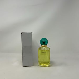 Könnte beinhalten: Eine Parfümflasche mit grünem Verschluss und goldenen Akzenten, neben einer grauen Schachtel. Die Flasche enthält eine gelbe Flüssigkeit und trägt die Aufschrift "Happy Chopard". Der Hintergrund ist schlicht weiß.