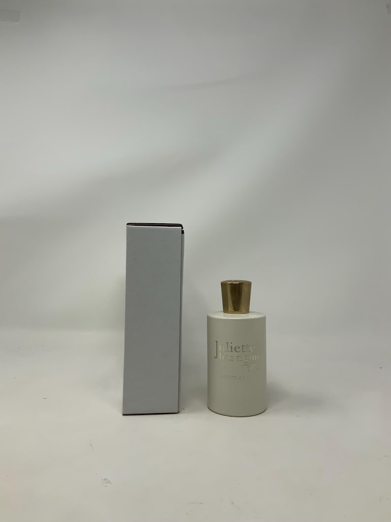 May include: A white perfume bottle with a gold cap and a matching rectangular box. The bottle has the text "Juliette Has A Gun" printed on it in a silver color. The box is a light gray color.