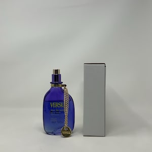 Könnte beinhalten: Eine blaue Parfümflasche mit goldenen Akzenten und einer goldenen Kette, neben einer weißen Schachtel. Auf der Flasche steht das Wort "VERSUS". Die Parfümflasche ist etwa 10 cm hoch.
