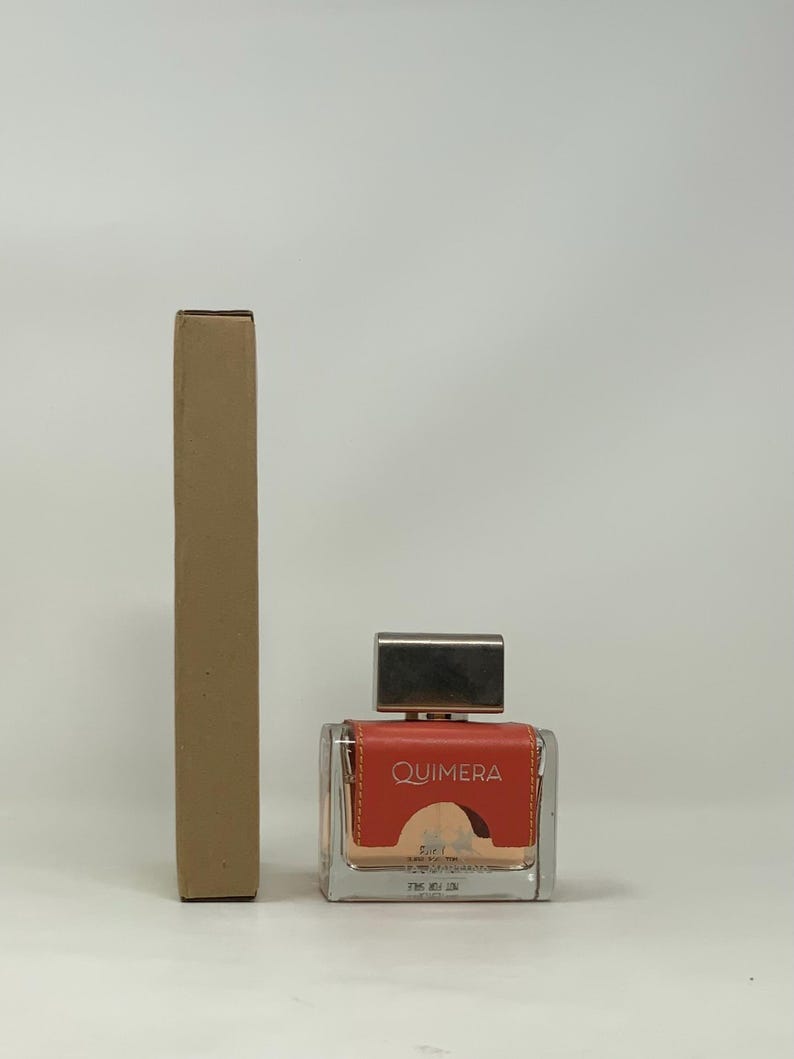 以下が含まれることがあります： 正方形の香水瓶と銀色のキャップ。ボトルには「QUIMERA」と印刷されたコーラル色のラベルが付いています。ボトルの隣には、背の高い長方形の段ボール箱があります。