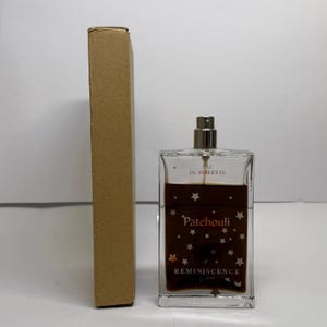 May include: A rectangular glass perfume bottle with a silver spray top, containing a brown liquid. The bottle is labelled "Patchouli" and "Reminiscence", decorated with star motifs. A brown cardboard box stands beside the bottle.