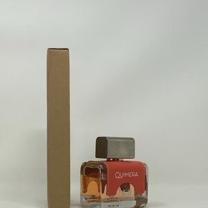 以下が含まれることがあります： 正方形の香水瓶と銀色のキャップ。ボトルには、白で「QUIMERA」という文字が入った赤いラベルが付いています。ボトルの左側には、長方形の段ボール箱があります。中の香水は明るいオレンジ色です。