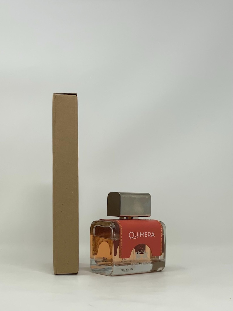 以下が含まれることがあります： 正方形の香水瓶と銀色のキャップ。ボトルには、白で「QUIMERA」という文字が入った赤いラベルが付いています。ボトルの左側には、長方形の段ボール箱があります。中の香水は明るいオレンジ色です。