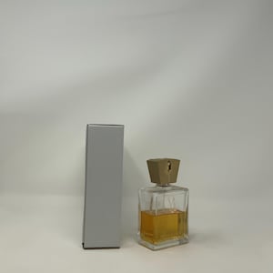 Puede incluir: Un frasco de perfume transparente y rectangular con un l&iacute;quido de color dorado en su interior y un tap&oacute;n geom&eacute;trico de color marr&oacute;n claro. El frasco est&aacute; junto a una caja gris, rectangular y alta. El frasco de perfume tiene la letra "V" en la parte delantera.
