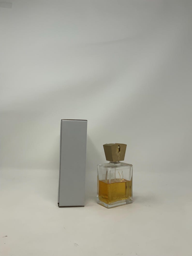 Puede incluir: Un frasco de perfume transparente y rectangular con un l&iacute;quido de color dorado en su interior y un tap&oacute;n geom&eacute;trico de color marr&oacute;n claro. El frasco est&aacute; junto a una caja gris, rectangular y alta. El frasco de perfume tiene la letra "V" en la parte delantera.