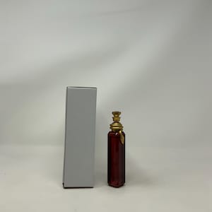 May include: A rectangular, dark red glass bottle with a gold-colored stopper and a white box. The bottle has a decorative design and is likely for perfume or a similar product. The stopper has a detailed, ornate design.