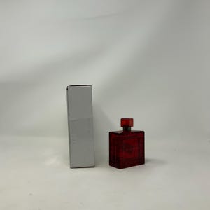 Könnte beinhalten: Eine rote Parfümflasche mit quadratischer Form und passendem quadratischem Deckel. Die Flasche steht neben einer hohen, rechteckigen, grauen Schachtel. Die Parfümflasche hat eine strukturierte Oberfläche und eine dunkelrote Farbe.