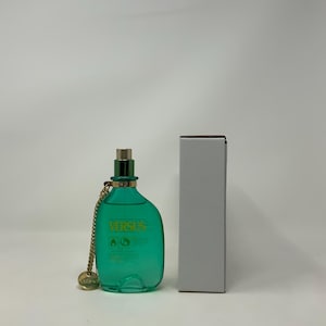 Könnte beinhalten: Eine türkisfarbene Parfümflasche mit goldener Kette und Verschluss. Auf der Flasche steht in Gelb "VERSUS". Daneben steht eine weiße rechteckige Schachtel.
