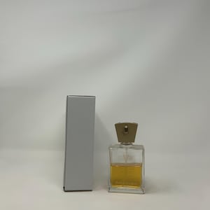 Puede incluir: Un frasco de perfume rectangular transparente con una tapa dorada y un l&iacute;quido &aacute;mbar claro en su interior. El frasco est&aacute; junto a una caja gris, rectangular y alta. El frasco de perfume tiene el nombre de la marca impreso.