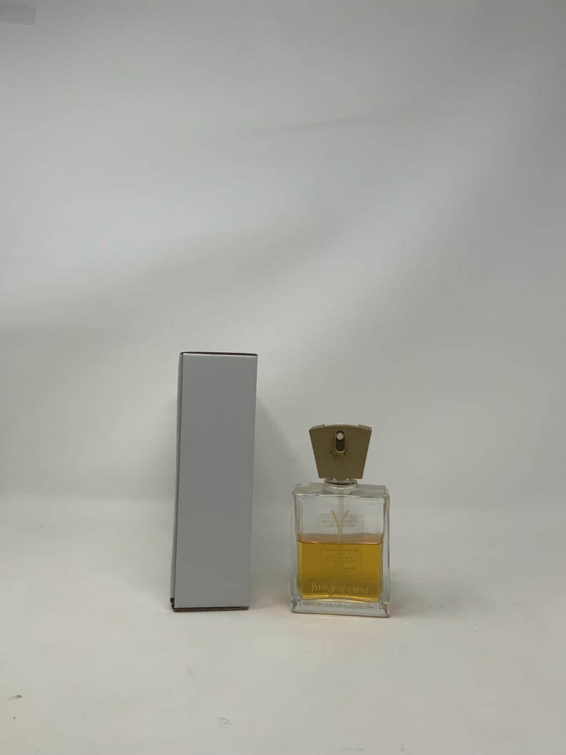 Puede incluir: Un frasco de perfume rectangular transparente con una tapa dorada y un l&iacute;quido &aacute;mbar claro en su interior. El frasco est&aacute; junto a una caja gris, rectangular y alta. El frasco de perfume tiene el nombre de la marca impreso.