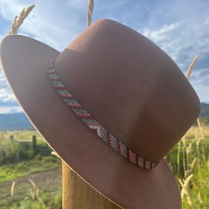 May include: A brown fedora-style hat with a decorative beaded band in red, white, and turquoise. The hat is set against a backdrop of a sunny sky and grassy field.