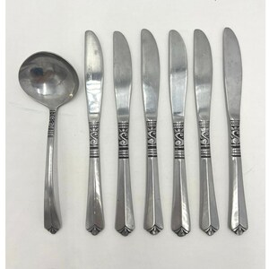 May include: A stainless steel flatware set comprising a soup ladle and six butter knives. The handles feature a decorative scroll pattern. The knives have a curved blade and a pointed tip. The set is presented against a white backdrop.