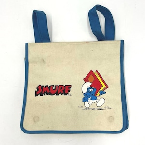 May include: Vintage Smurf tote bag with blue trim and handles. The bag features the word "SMURF" in red and a cartoon Smurf character carrying a book. The bag is made of a beige fabric.