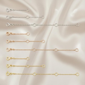 May include: A collection of jewelry chain extenders in silver, rose gold, and gold tones. Each extender features a lobster clasp on one end, a small ring in the middle, and a larger ring on the other end. The extenders are displayed on a cream-colored fabric.