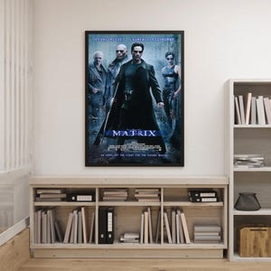 Może przedstawiać: Oprawiony plakat filmowy do filmu "Matrix" z Keanu Reevesem, Laurence'em Fishburne'em i Carrie-Anne Moss. Plakat ma ciemną kolorystykę z tytułem "MATRIX" dużymi, pogrubionymi literami. Plakat jest wyświetlany nad drewnianą półką wypełnioną książkami i segregatorami.