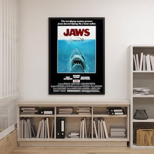 Op de afbeelding: Een ingelijste filmposter voor de film "Jaws" met een haai die uit blauw water opduikt. De poster heeft het woord "JAWS" in grote witte letters, met de namen van de acteurs en de slogan "De angstaanjagende film van de angstaanjagende nr. 1 bestseller".