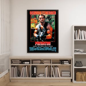 Puede incluir: Un póster enmarcado de la película "Predator" con Arnold Schwarzenegger. El póster tiene un borde negro con texto rojo. La imagen muestra a Schwarzenegger sosteniendo un arma, con un fondo de selva. El texto "SOON THE HUNT WILL BEGIN" también es visible.