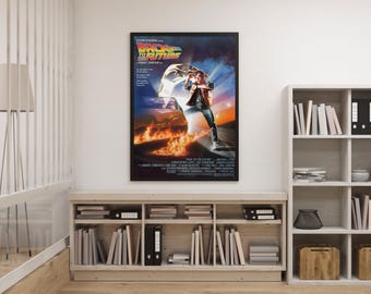 May include: A framed movie poster for "Back to the Future" hangs on a white wall. The poster features a dynamic image of a man in a vest and jeans, with a car engulfed in flames. The title is in bold, colorful text.