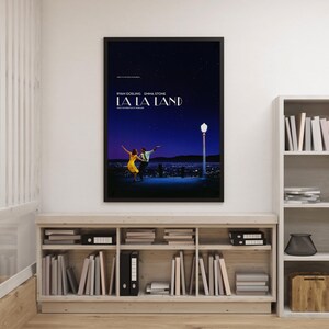 Może przedstawiać: Oprawiony plakat filmowy do filmu "La La Land" z Ryanem Goslingiem i Emmą Stone. Plakat przedstawia parę tańczącą pod gwiaździstym niebem, z światłami miasta w tle. Plakat znajduje się na jasnej drewnianej półce z książkami.