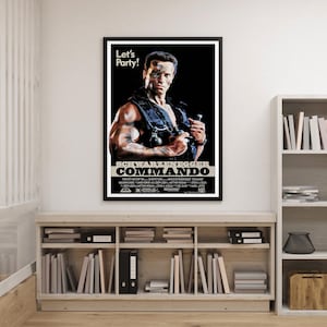 Puede incluir: Un póster enmarcado de la película "Commando" con Arnold Schwarzenegger. El póster tiene un fondo negro con el título de la película y la frase "Let's Party!" en la parte superior. El póster se muestra en una estantería de madera clara.