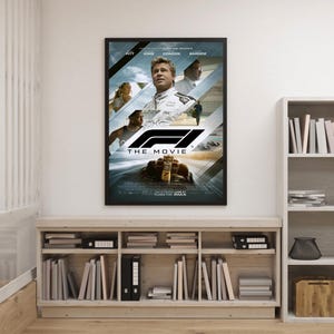 May include: Framed movie poster for "F1: The Movie" featuring Brad Pitt. The poster has a black frame and showcases a dynamic image of a Formula 1 race car and drivers. The poster is displayed above a wooden bookshelf filled with books and office supplies.