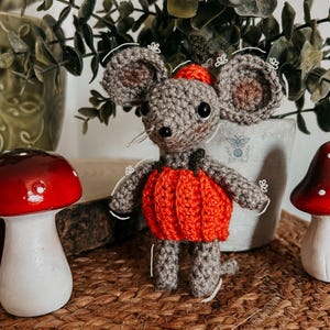 May include: A crocheted gray mouse wearing an orange pumpkin costume and a small red hat. The mouse has large ears and a long tail. Two decorative red and white mushroom figurines are on either side of the mouse.