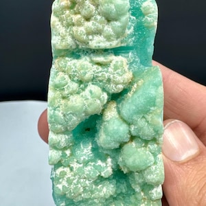 May include: A vibrant, textured mineral specimen with a light teal and white color palette. The surface has a bubbly, layered appearance. The stone is held in a hand, showcasing its natural, irregular shape. The overall aesthetic suggests a raw, natural gemstone.