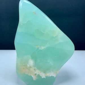 May include: A polished, irregular-shaped piece of mint green stone with translucent qualities. The surface displays smooth, glossy areas interspersed with cloudy, opaque white and pale yellow mineral inclusions throughout the structure.