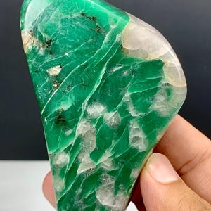 Op de afbeelding: Een gepolijste, onregelmatig gevormde groene chrysopraassteen met doorschijnende witte kwartsinsluitingen en kleine donkere mineraalvlekjes. Het oppervlak heeft een gladde, glanzende textuur met zichtbare interne breuklijnen en lagen.