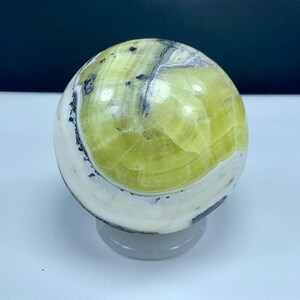May include: A polished spherical stone featuring a smooth surface with prominent bands of lime green, creamy white, and dark gray mineral inclusions. The sphere displays natural concentric patterns and sits on a clear circular stand.