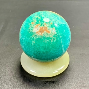 May include: A vibrant turquoise sphere with brown and white markings sits atop a pale yellow, circular base. The sphere has a polished, smooth surface. The base is a solid, opaque color. The object is displayed on a dark surface.