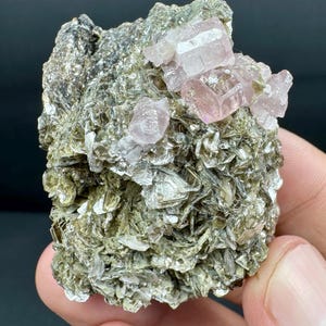 May include: A natural mineral specimen featuring a cluster of pale pink crystals atop a matrix of silvery-green, layered mineral flakes. The crystals have a translucent appearance, and the overall texture is rough and uneven.