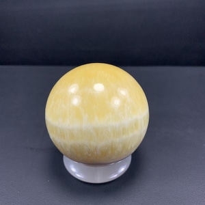 May include: A polished, yellow sphere with white banding, resting on a small, white, circular stand. The gemstone ball has a smooth, reflective surface. The background is a dark gray color.