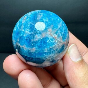 May include: A polished, spherical gemstone in shades of blue, with a white inclusion. The stone has a translucent quality, revealing internal patterns and textures. The sphere is held in a hand against a dark background.