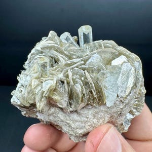May include: A natural mineral specimen with light green mica flakes and clear, elongated aquamarine crystals. The base is a rough, light grey rock. The overall appearance is crystalline and textured.