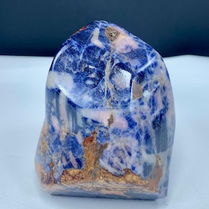 Op de afbeelding: Een gepolijste sodalietsteen met een diepblauw en wit gemarmerd patroon met duidelijke vlekken van lichtbruine en bruine minerale insluitingen. De steen heeft een gladde, afgeronde bovenkant en een natuurlijke, onregelmatige basis met scheuren.