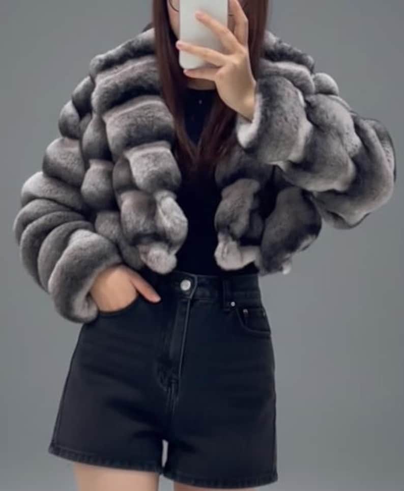 A person is taking a selfie with a grey and black fur coat, black shorts, and a black top.
