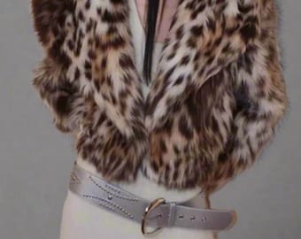 May include: A cropped faux fur jacket with a leopard print pattern in shades of brown and tan. The jacket has a wide collar and is paired with a silver belt with studded details. The outfit is completed with a cream-colored dress.