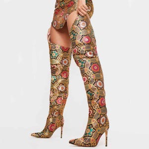 May include: Over-the-knee boots with a pointed toe and stiletto heel. The boots feature a gold, brown, pink, and teal floral pattern. The boots are made of a shiny material and have a high heel.