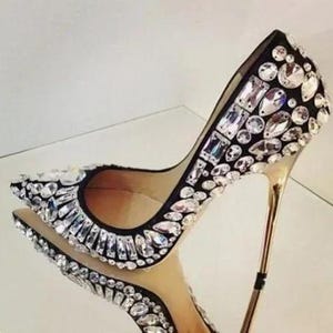 May include: Black high-heeled shoe embellished with clear rhinestones. The stiletto heel is gold-colored. The shoe is reflected in a mirrored surface. The rhinestones are various shapes and sizes, adding sparkle and glamour to the footwear.