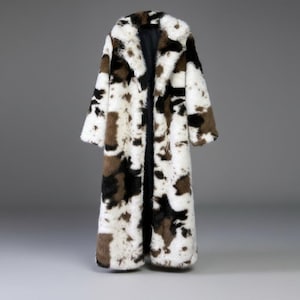 Peut inclure: Long manteau en fausse fourrure avec un motif de peau de vache. Le manteau a un col cranté et des manches longues. La palette de couleurs comprend des taches blanches, marron et noires, créant un contraste saisissant. Le manteau est ouvert à l'avant.