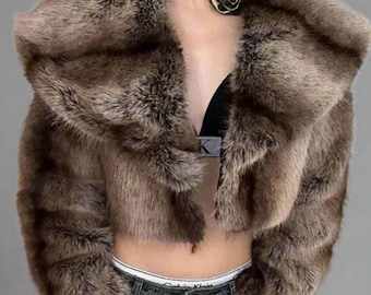 May include: A cropped brown faux fur jacket with a wide collar. The jacket has a textured appearance and a dark brown color. The model is wearing blue jeans and a black bra. A gold-colored rose pendant is worn around the neck.