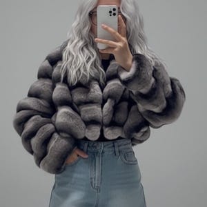 A person with long, wavy hair is taking a selfie with a smartphone, wearing a gray fur coat and denim shorts.