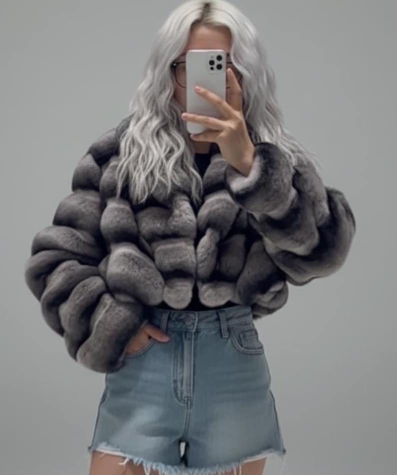 A person with long, wavy hair is taking a selfie with a smartphone, wearing a gray fur coat and denim shorts.