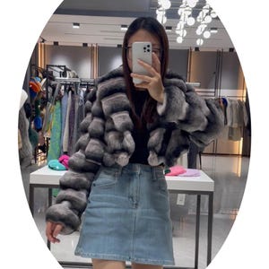 A woman wearing a gray fur coat and denim skirt is taking a selfie in a clothing store.