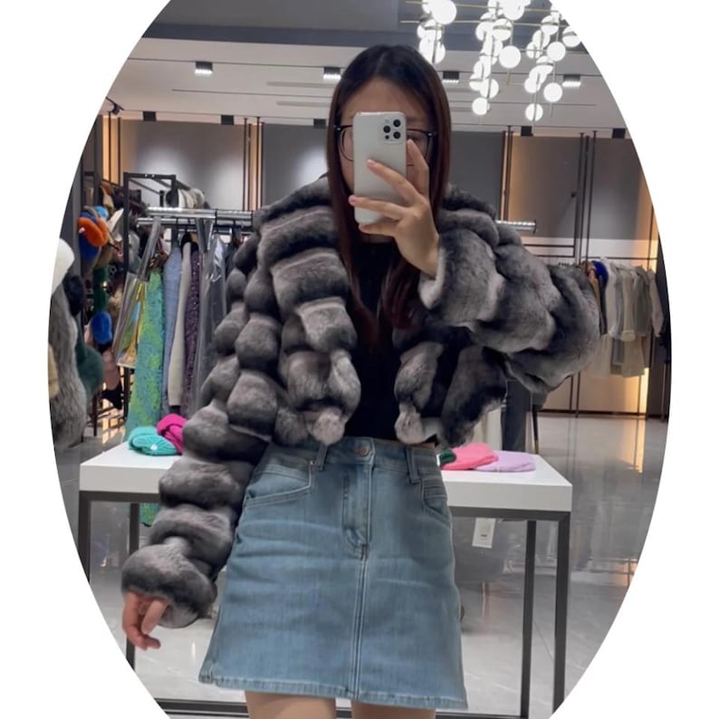 A woman wearing a gray fur coat and denim skirt is taking a selfie in a clothing store.