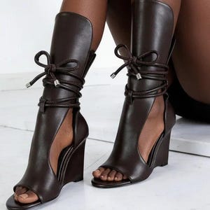 Puede incluir: Sandalias de cuña marrón oscuro con punta abierta y caña alta. Las sandalias presentan un corte lateral y están atadas con un material similar a una cuerda, atado con un lazo en la parte superior. El tacón de cuña mide aproximadamente 10 cm de alto.