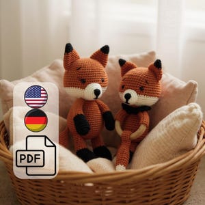 Könnte beinhalten: Zwei handgefertigte Fuchs-Puppen aus Häkelarbeit in einem Weidenkorb. Die Füchse sind orange mit weißen und schwarzen Akzenten. Die Puppen haben schwarze Augen und liegen auf einem beigen Kissen. Das Bild enthält ein PDF-Symbol und die Flaggen der USA und Deutschlands.